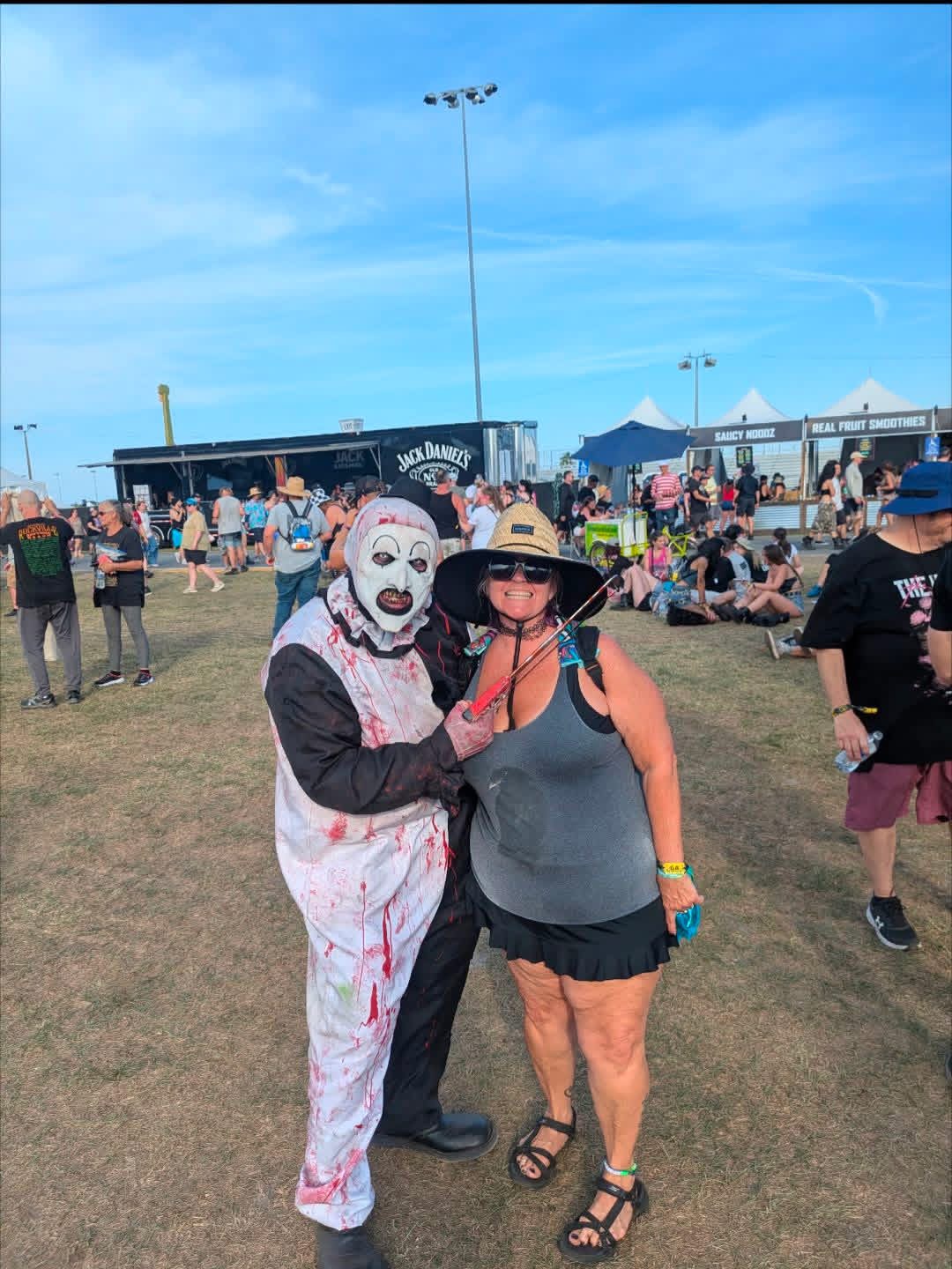 Art the Clown posing with a fan in a straw hat by the food trucks on a blue-sky day
