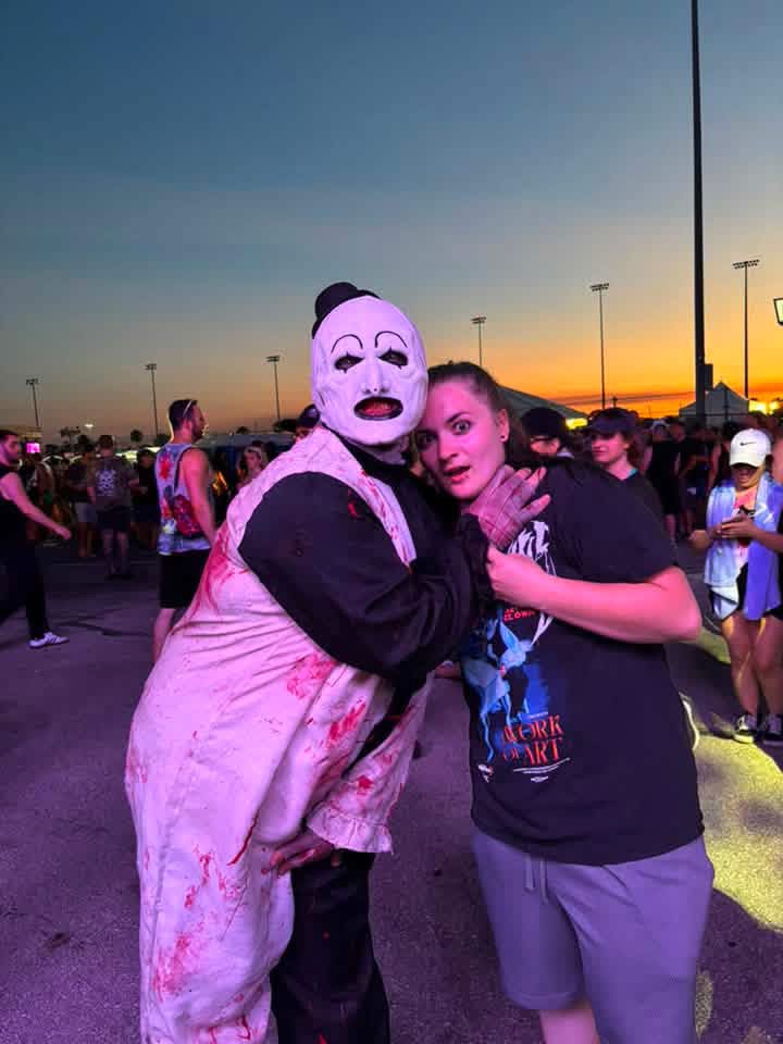 Art the Clown posing with a fan under an orange sunset in the festival lot
