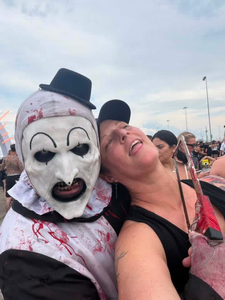 Art the Clown leaning cheek to cheek with a laughing fan in a cap